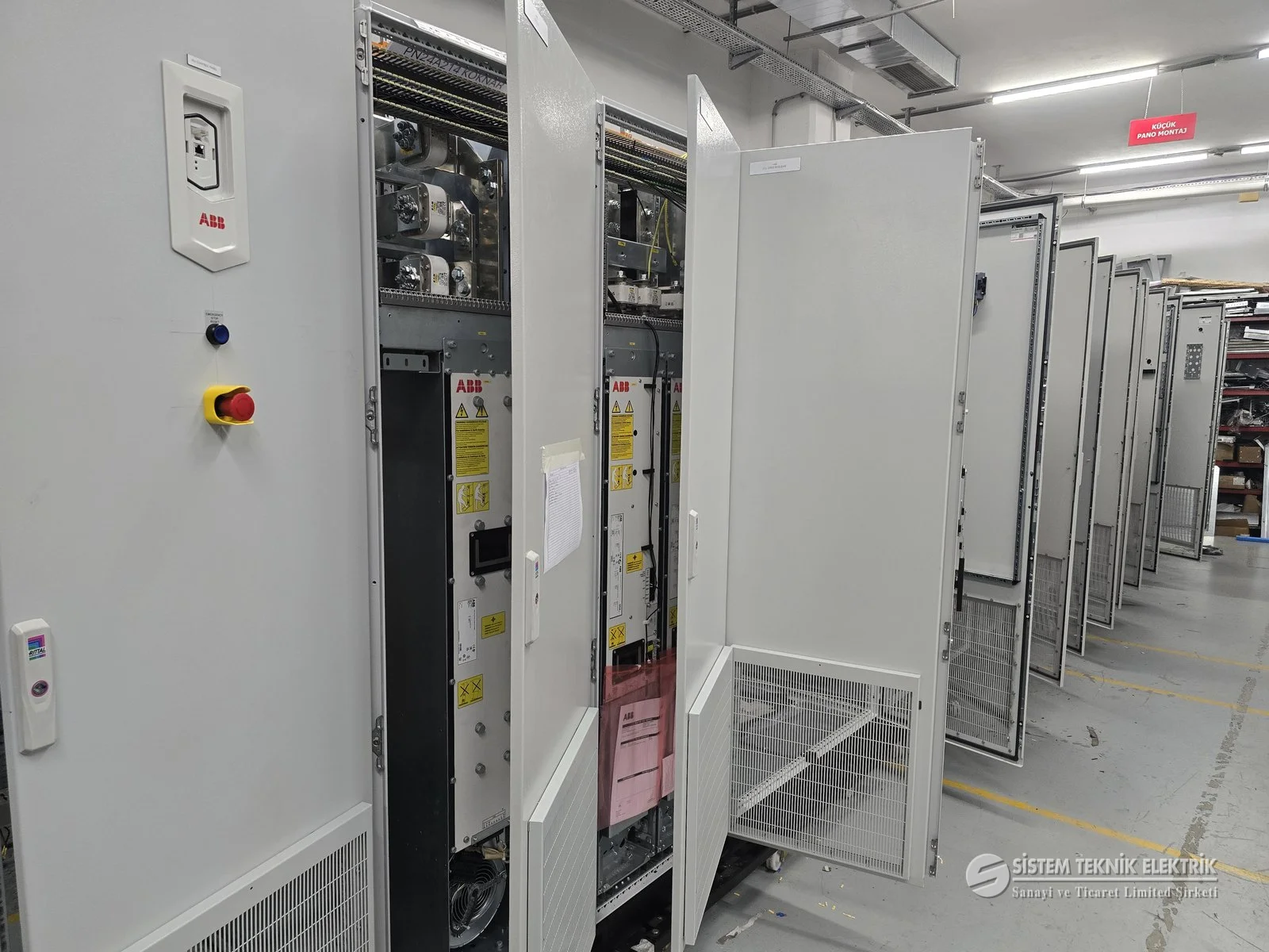 Pano İmalatı ve Elektrik Projelendirme — Açık kapaklı pano, arkada uzun pano dizisi.