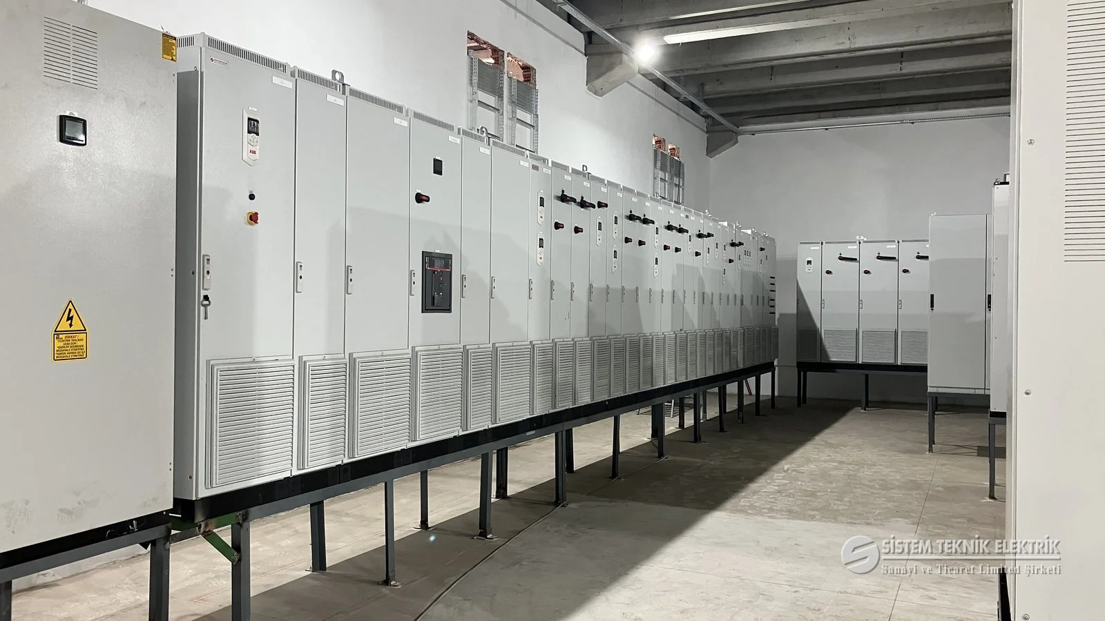 Pano İmalatı ve Elektrik Projelendirme — kaide üstünde uzun gri pano dizisi, perspektif.